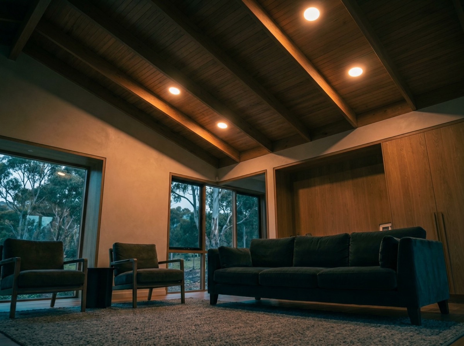 A living room with downlights on in a raked timber ceiling and gum trees through the windows