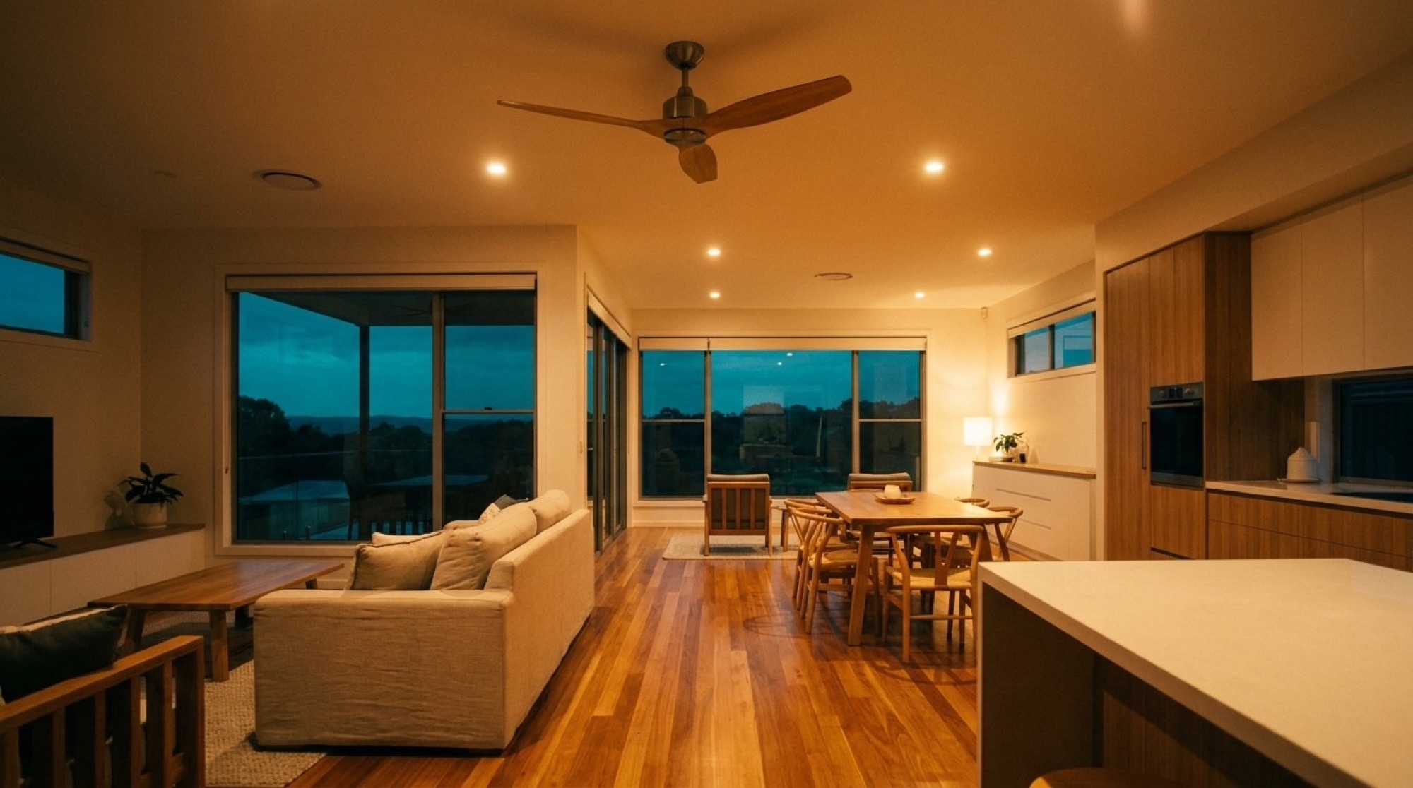 A living area at dusk with a ceiling fan and downlights on above a timber floor