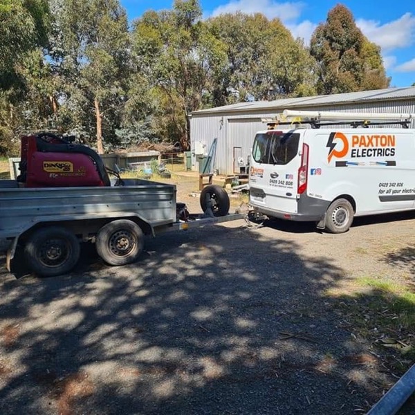 Two Paxton Electrics vans and a work trailer parked on a gravel driveway among gum trees